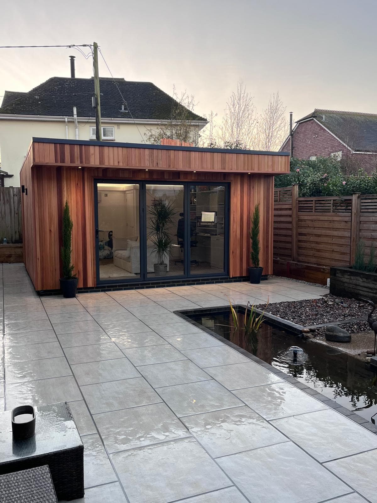 Cedar-clad garden room with bi-fold doors beside a landscaped patio and pond, built by FR Carpentry
